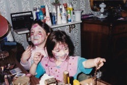 Two little girls, all the skin on their faces covered in a variety of different colored make-up, sitting at a make-up vanity.